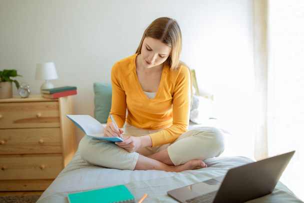 Eラーニング。若い大学生の女性は居心地の良いベッドルームのインテリアでベッドの上にノートパソコンの近くに座って教科書でノートを取ります.十代の女性は自宅で大学の宿題をしている。距離研究 - 写真・画像