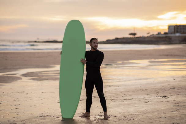 Bonito surfista homem com sua prancha de surf em pé na praia ao pôr do sol, atlético jovem masculino em roupa de mergulho relaxante após o surf, desfrutando de esportes aquáticos e estilo de vida ativo, espaço de cópia - Foto, Imagem