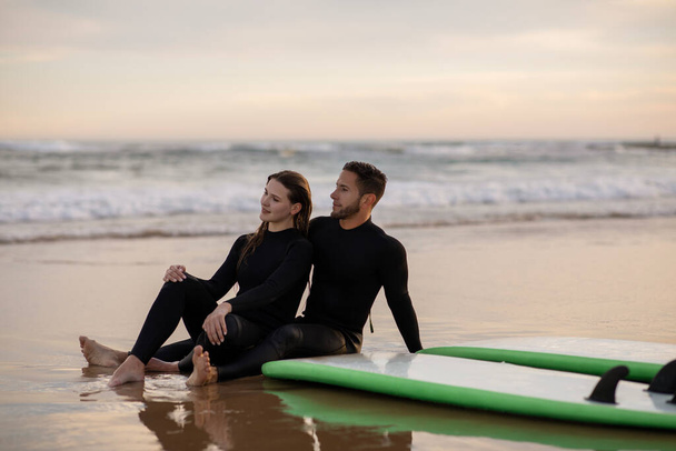Happy young couple chilling out on the beach after surfing, millennial surfers man and woman wearing wetsuits sitting on sand near their boards, romantic lovers looking at sunset, copy space - Photo, image