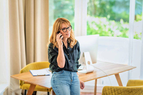 Shot of mid aged woman standing at desk and using mobile phone. Confident female wearing glasses and casual clothes and having a call. Home office. - Valokuva, kuva