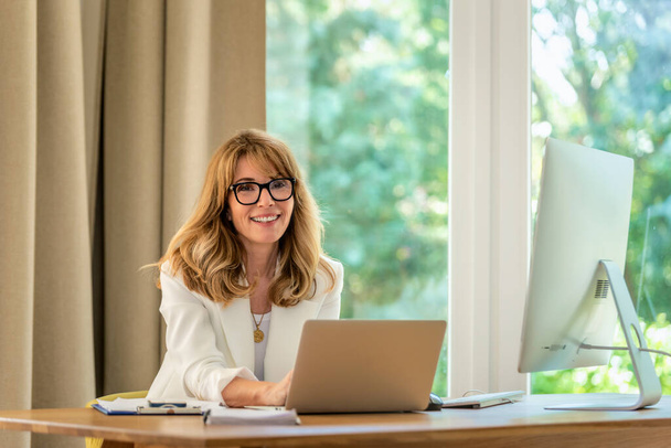 Cheerful smiling mid aged businesswoman sitting at desk and using laptop for work. Confident professional female wearing white blazer and eyewear.  - Valokuva, kuva