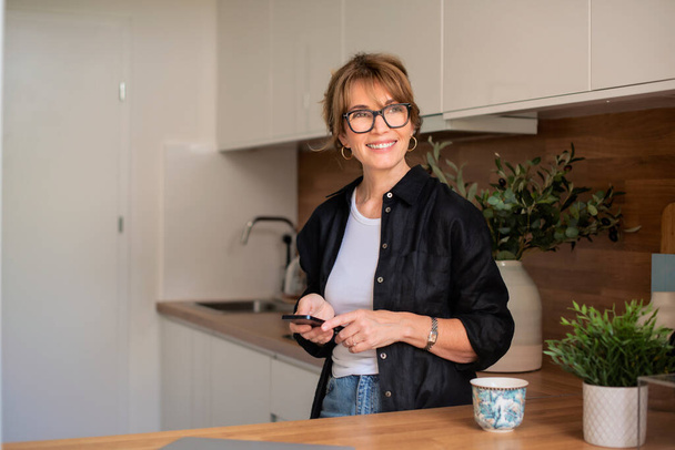 Shot of middle aged woman with mobile phone standing in the kitchen at home. Home office. - Valokuva, kuva