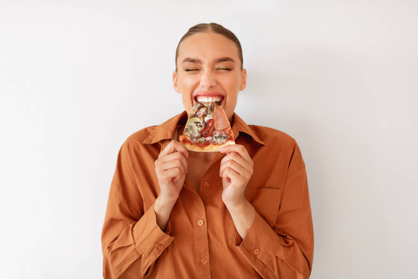 Donna eccitata godendo pizza holding e mordere gustosa fetta in piedi su sfondo muro bianco. Amante del cibo spazzatura mangiare pizza italiana. Nutrizione malsana cheat pasto - Foto, immagini