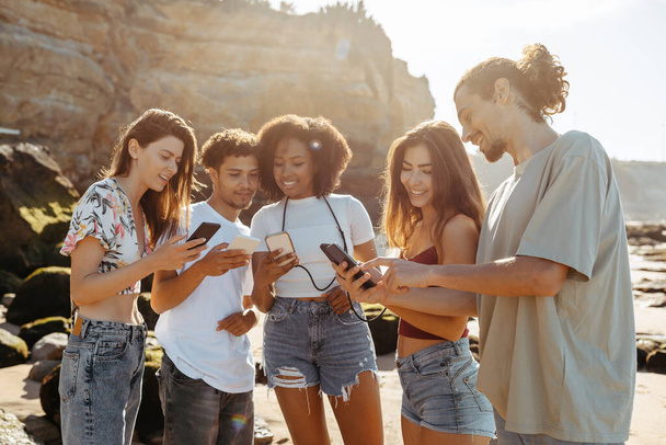 Sorrindo millennial multirracial pessoas estudante desfrutar de férias de verão, conversando em redes sociais em telefones na praia. Jogo, site recomendação, blog, app e estilo de vida moderno, fim de semana juntos - Foto, Imagem