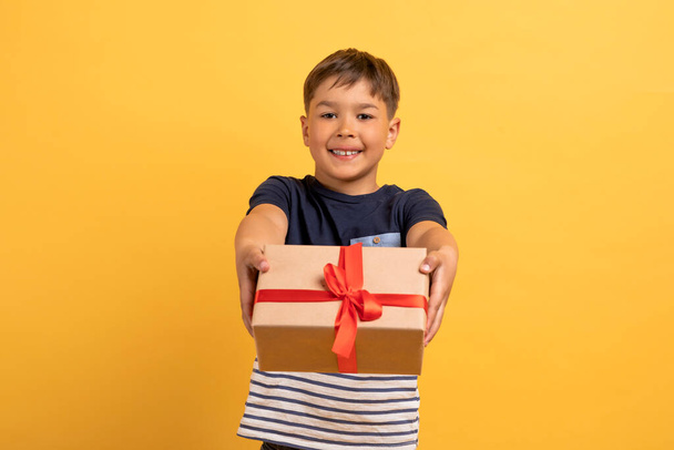 Portret uroczego, słodkiego chłopca w wieku szkolnym trzymającego pudełko prezentów i patrzącego na odizolowaną kamerę na żółtym tle studia. Dziecko na przyjęciu urodzinowym, przestrzeń do kopiowania - Zdjęcie, obraz