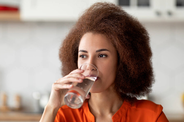 Gezond vloeibaar. Mooie zwarte vrouw drinken mineraalwater uit glas in keuken, dorstige jonge Afro-Amerikaanse dame genieten van verfrissend drankje thuis, close-up portret, vrije ruimte - Foto, afbeelding