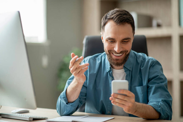 Усміхнений красивий молодий бізнесмен перевіряє бізнес мобільний додаток під час роботи в сучасному офісі, щасливий тисячолітній чоловік-підприємець сидить на робочому столі і використовує смартфон, копіює простір
 - Фото, зображення