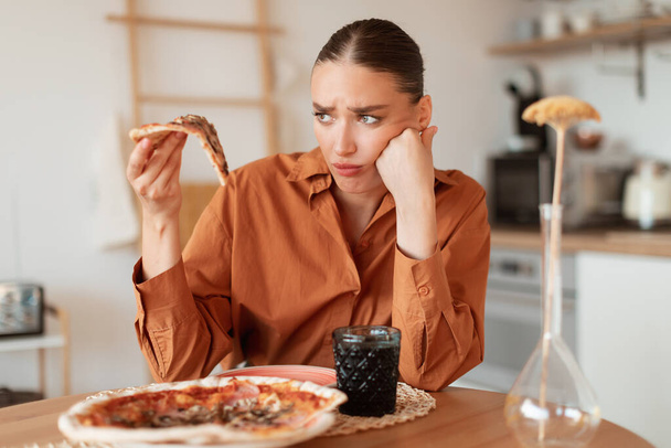 Donna sconvolta guardando delizioso pezzo di pizza ad alto contenuto calorico, tenendo fetta in mano. Dieta e problemi digestivi, nonché cibo viziato e insipido - Foto, immagini