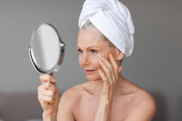 Eye wrinkles treatment. Beautiful european mature lady using moisturizing eye cream, looking at mirror, enjoying face care routine, closeup shot - Photo, Image