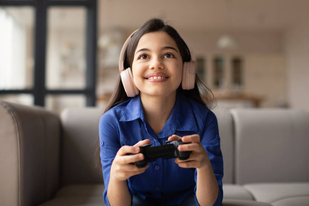 Happy Arabic Kid Girl Playing Video Game With Gaming Console Sitting On Couch At Home On Weekend. Preteen Child Enjoying Videogame Winning And Having Fun. Digital Leisure And Entertainment - Foto, immagini