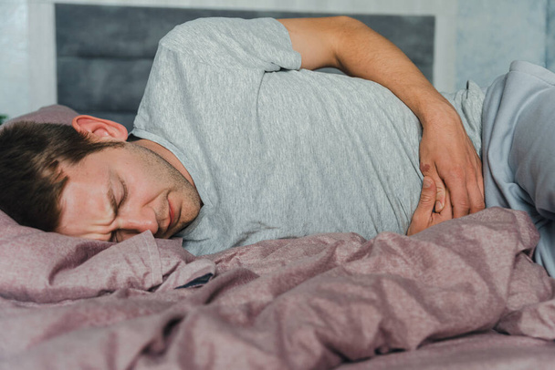 Frustrated handsome young man hugging his belly suffering from stomach pain while lying in bad at home. Male with belly ache. Stomach ulcer, gastritis, food poisoning concept - Photo, Image