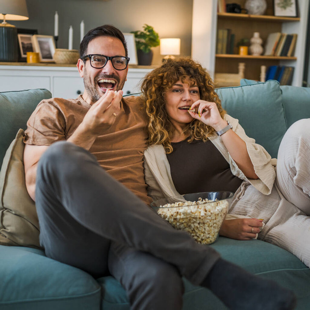 Couple man and woman caucasian husband and wife or boyfriend and girlfriend sit on the sofa bed at home watch tv movie series with bowl of popcorn happy smile bonding love family concept copy space - Foto, Imagen