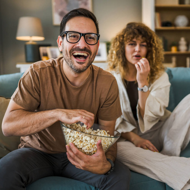 Couple man and woman caucasian husband and wife or boyfriend and girlfriend sit on the sofa bed at home watch tv movie series with bowl of popcorn happy smile bonding love family concept copy space - Фото, зображення