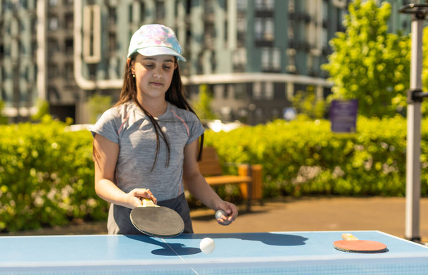 Young teenager girl playing ping pong. She holds a ball and a racket in her hands. Playing table tennis outdoors in the yard. - Foto, Bild