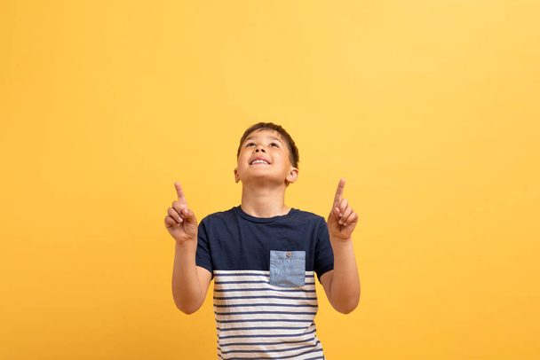 Cool cheerful boy schooler in casual t-shirt pointing up at copy space for text or advertisement above his head and smiling, isolated on yellow studio bakground. Great offer for kids - Fotografie, Obrázek