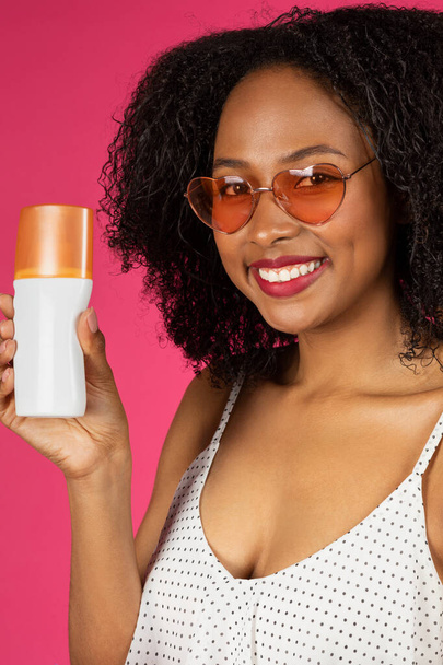 Happy millennial black woman in sunglasses show jar of sun cream, enjoy summer vacation, isolated on magenta studio background, close up, vertical. Skin protection recommendation, cosmetics - Photo, Image