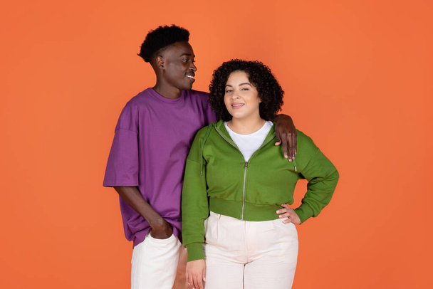 Loving happy beautiful millennial multicultural couple handsome black guy and pretty chubby curly brunette woman wearing casual outfit posing on orange studio background - Foto, immagini