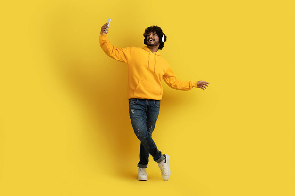 Cheerful joyful young stylish handsome indian guy wearing casual outfit taking selfie on smartphone while listening to music over yellow background, using wireless stereo headphones, copy space - Fotografie, Obrázek