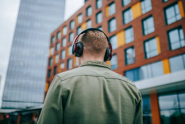 A male with earbuds in his ears and his back facing the camera, experiencing the city vibes while listening to music. Male teenager with headphones listening to music, rear view - Foto, Bild