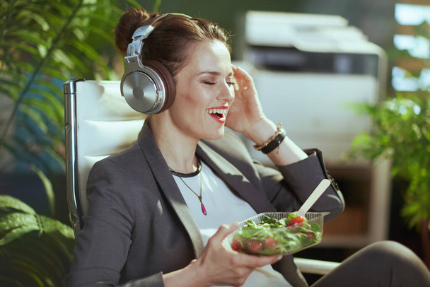 Βιώσιμος χώρος εργασίας χαρούμενη μοντέρνα μεσήλικη λογίστρια με γκρι κοστούμι σε μοντέρνο πράσινο γραφείο με ακουστικά που τρώει σαλάτα. - Φωτογραφία, εικόνα