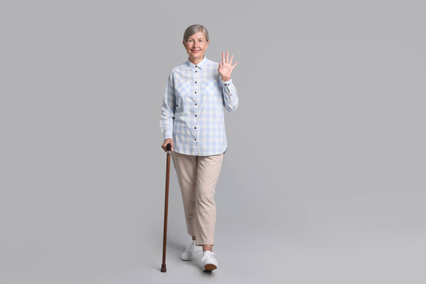 Senior woman with walking cane waving on gray background - Fotó, kép