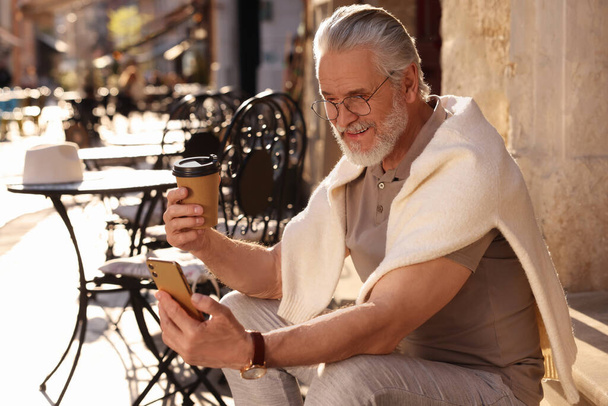 Handsome senior man sitting on doorstep, using smartphone and drinking coffee outdoors, space for text - Φωτογραφία, εικόνα