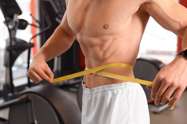 Athletic man measuring waist with tape in gym, closeup - Foto, Imagen