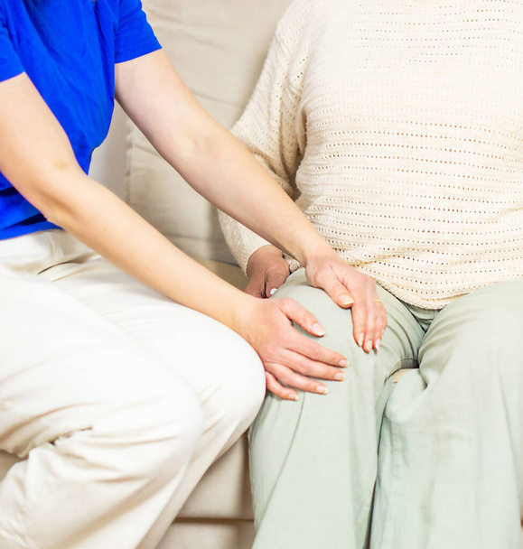 Młoda profesjonalna opiekunka pomaga starszej kobiecie z bólem kolana w domu. Przyjazna pielęgniarka asystująca pacjentce w średnim wieku. Emerytowana staruszka siedzi na kanapie i gada - Zdjęcie, obraz