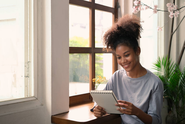 Freelancer feminino afro-americano satisfeito sentado em casa aconchegante interior e verificando o plano de trabalho no planejador diário pessoal. - Foto, Imagem