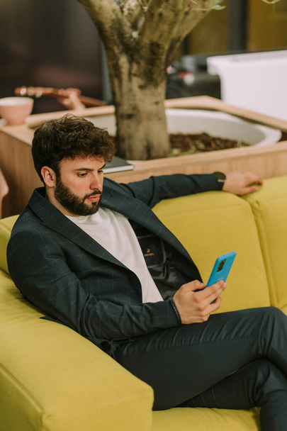 Good looking male business person checking his phone while sitting on yellow sofa at the office after work - Foto, Bild