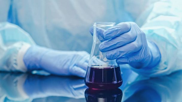 Laboratory research. Drugs development. Medical chemistry. Technician in protective gloves checking studying reaction in filled glass flask in hands. - Photo, Image