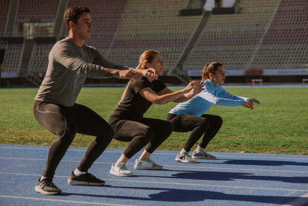 Ein Mann und zwei Frauen trainieren gemeinsam im Stadion. Sie machen Kniebeugen - Foto, Bild