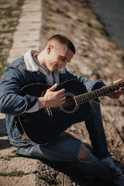 Κοντινό πλάνο του όμορφου άντρα που παίζει κιθάρα ενώ κάθεται στο έδαφος δίπλα στο ποτάμι - Φωτογραφία, εικόνα