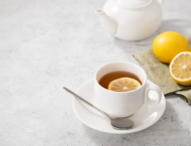 A  cup of tea with lemon and spoon on a light background with a white teapot and citrus. Healthy morning drink concept. Free space for text. - Photo, Image
