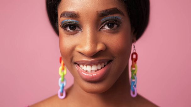 Color makeup. Happy face. Summer beauty. Cheerful smiling woman with full lips blue eye shadow long eyelashes rainbow earrings on pink free space. - Foto, imagen
