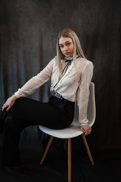  portrait of beautiful blonde woman in white blouse sitting on chair isolated on black, studio - 写真・画像