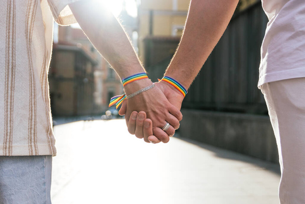 Close-up photo of two unidentified men's hands, tightly holding each other while walking along a sun-drenched city street at sunset. They wear rainbow-colored LGBT bracelets on their wrists. - Photo, Image