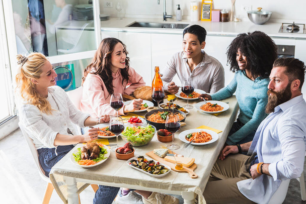 Grupo de amigos multiétnicos sentados a la mesa durante el almuerzo. Mesa con espaguetis con salsa de tomate, pollo asado, verduras y copas de vino tinto. Ambiente de alegría - Foto, Imagen