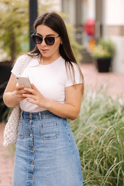 Pretty woman in denim skirt and white t-shirt using phone. Woman walking outdoors and looking for something in social networks. High quality photo - Zdjęcie, obraz