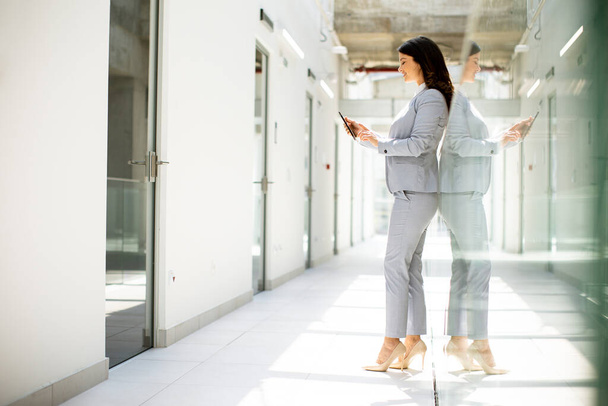 Красивая молодая деловая женщина пользуется мобильным телефоном в офисном коридоре - Фото, изображение