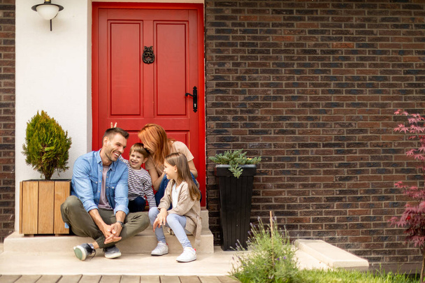 Gezin met een moeder, vader, zoon en dochter zitten buiten op stappen van een veranda van een bakstenen huis - Foto, afbeelding