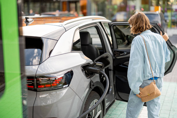 Jeune femme ouvre la porte du véhicule de sa voiture électrique, la rechargeant sur une borne de recharge publique - Photo, image