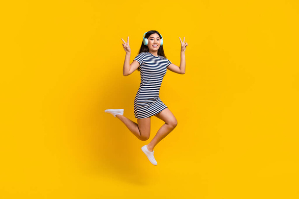 Full length photo of cheerful pretty lady wear striped dress earphones jumping showing v-signs isolated yellow color background. - Φωτογραφία, εικόνα