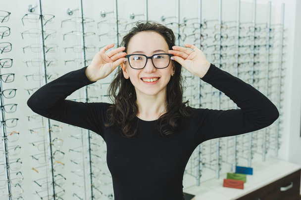 Egy csinos nő szemüveget választ egy szemészeti boltban. Egy nő szemüveget vesz. Egy nő a háttérben a kirakatok különböző modellek szemüveg. - Fotó, kép