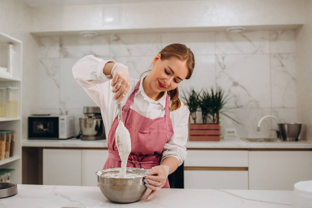 Portrait of confectioner woman In apron preparing to cook. Young girl whipping eggs in bow. Housewife is standing in the kitchen with a whisk. Young woman making whipped cream. Delicious food concept - Фото, зображення