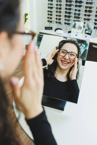 Optics shop. A young woman chooses glasses in an optical store. Stand with glasses. Vision correction. Woman in glasses. Portrait of a woman in corrective glasses. - Foto, Imagem