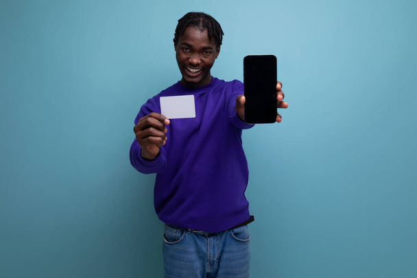 Αμερικανός νεαρός μελαχρινός άνδρας με dreadlocks κρατώντας κάρτα και smartphone mockup για ψώνια. - Φωτογραφία, εικόνα