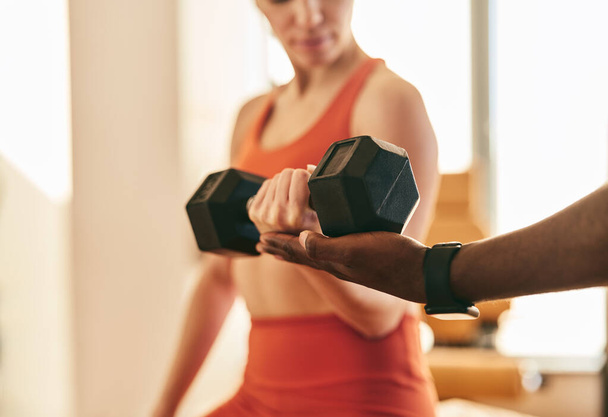 Crop anonymous female in red activewear lifting heavy dumbbells while working out in modern gym, being helped by instructor hand - 写真・画像