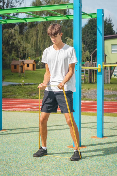 Goal-oriented teenager working on his body using resistance rubber performs strength training. Preparing for the sports season. Exercising with your own body. Outdoor workout playground. - Фото, изображение