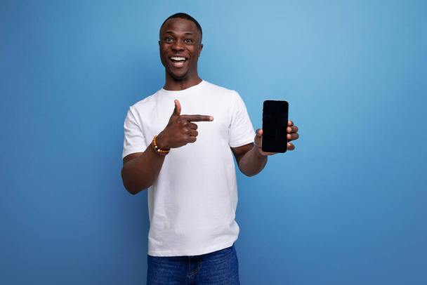 白いTシャツの素敵な若いアフリカの男はモックアップでスマートフォンを示しています. - 写真・画像
